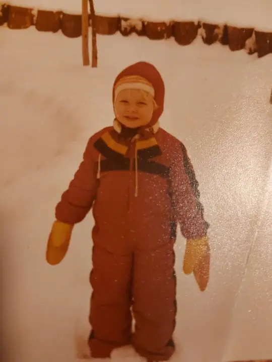 A small child in a red snowsuit, knit cap and big yellow mittens standing in deep snow