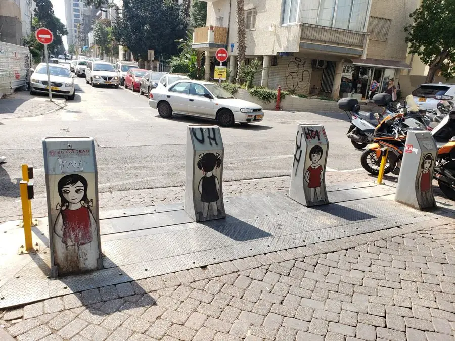 Four metal street bollards each painted with a girl in a red dress at a sunny street corner in Israel