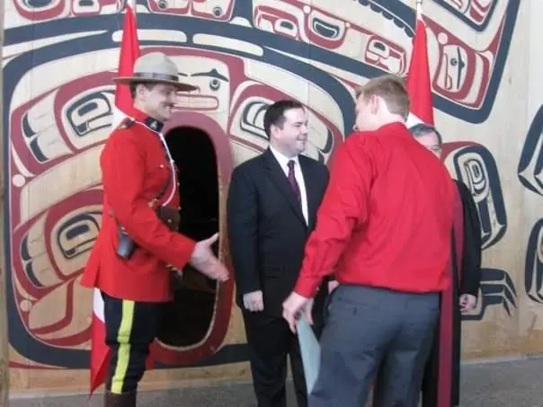 A citizenship ceremony in Ottawa: a Mountie in red serge, an official in a suit, and a man in a red shirt shaking hands in front of a carved Pacific Northwest wall design