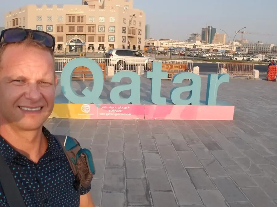 A smiling man taking a selfie in front of large teal 3D letters spelling Qatar on a paved plaza in Doha