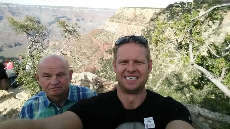 A smiling man in a black T-shirt and an older man in a plaid shirt taking a selfie at an overlook above the Grand Canyon