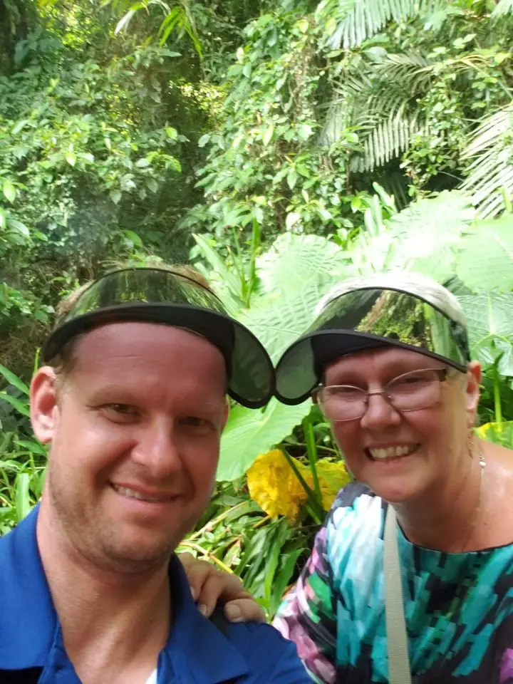 A smiling man and his mother wearing matching dark sun visors, taking a selfie among lush green tropical plants