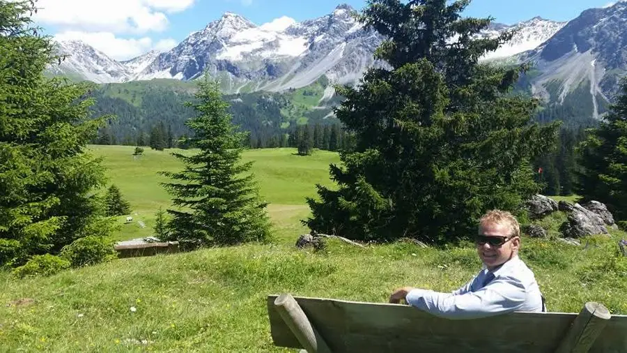 Me on a wooden bench in an alpine meadow with snowy peaks behind