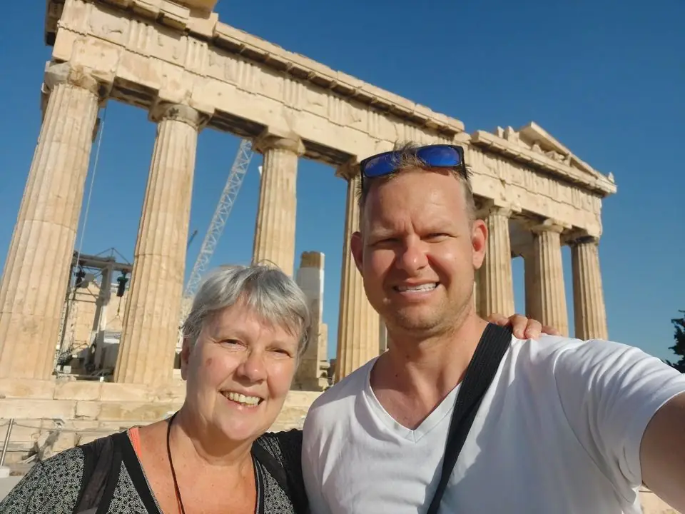 A smiling man and his mother taking a selfie in front of the Parthenon in Athens under a deep blue sky